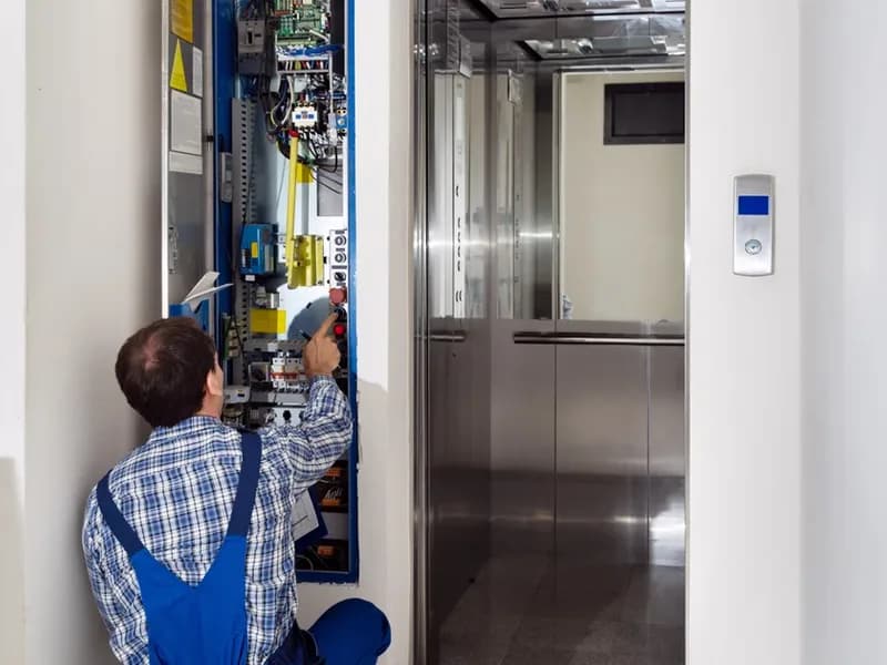 Technik v montérkách pracuje na rozvaděči výtahu vedle otevřené kabiny v chodbě domu