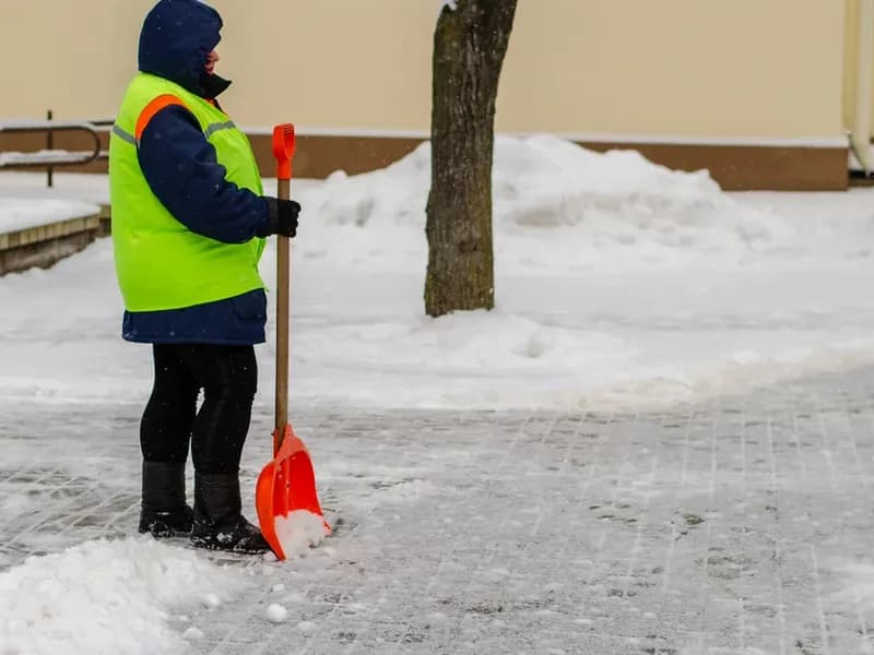 Pracovník v reflexní vestě odhrnuje lopatou sníh z chodníku před bytovým domem